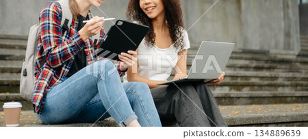 Three young college students is reading a book while relaxing sitting on grass in a campus park with her friends. 134889639