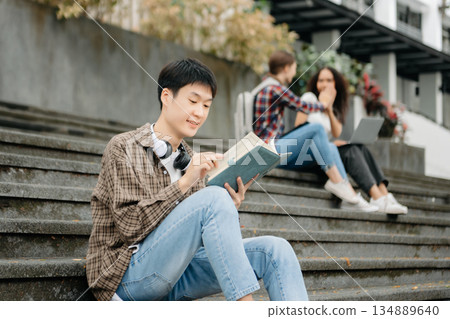 Three young college students is reading a book while relaxing sitting on grass in a campus park with her friends. 134889640