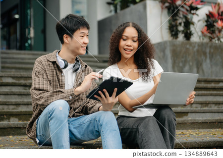Three young college students is reading a book while relaxing sitting on grass in a campus park with her friends. Three young college students is reading a book while relaxing sitting on grass in a campus park with her friends. 134889643