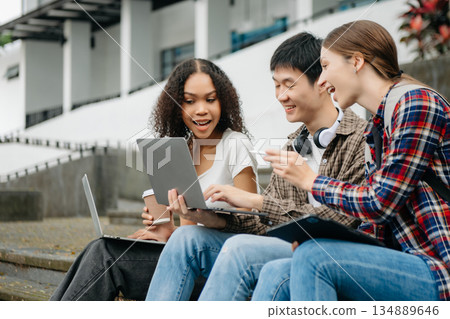 Three young college students is reading a book while relaxing sitting on grass in a campus park with her friends. Three young college students is reading a book while relaxing sitting on grass in a campus park with her friends. 134889646