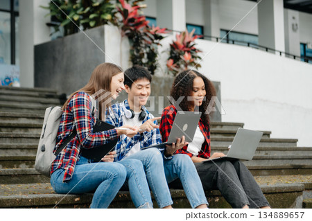 Three young college students is reading a book while relaxing sitting on grass in a campus park with her friends. 134889657