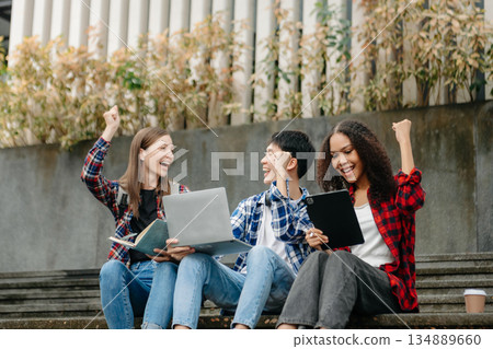 Three young college students is reading a book while relaxing sitting on grass in a campus park with her friends. 134889660