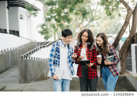 Three young college students is reading a book while relaxing sitting on grass in a campus park with her friends. 134889664