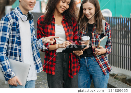 Three young college students is reading a book while relaxing sitting on grass in a campus park with her friends. 134889665