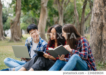 Three young college students is reading a book while relaxing sitting on grass in a campus park with her friends. 134889694