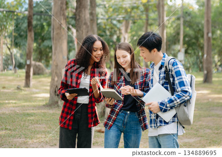 Three young college students is reading a book while relaxing sitting on grass in a campus park with her friends. Three young college students is reading a book while relaxing sitting on grass in a campus park with her friends. 134889695