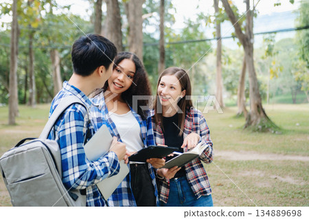 Three young college students is reading a book while relaxing sitting on grass in a campus park with her friends. 134889698