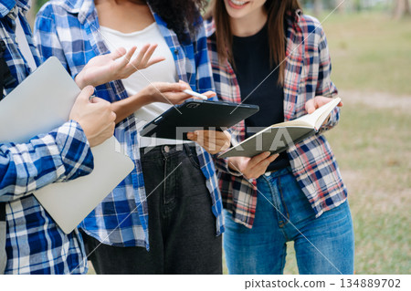 Three young college students is reading a book while relaxing sitting on grass in a campus park with her friends. 134889702