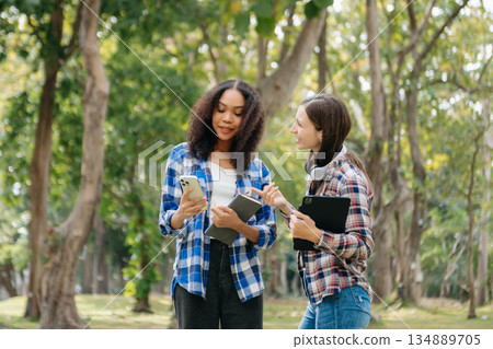 Three young college students is reading a book while relaxing sitting on grass in a campus park with her friends. 134889705