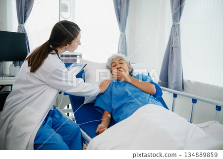 Doctor in white suit take notes while discussing and Asian elderly, woman patient who lying on bed with receiving saline solution in hospital or clinic. 134889859