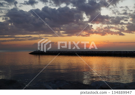 The first rays of sun paint the gray sky orange. The gray and orange sky is reflected in the sea, against the backdrop of a stone pier and a blinking lighthouse. 134889947