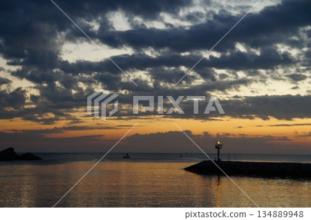 The dawn is breaking over the horizon.The first rays of sun paint the gray sky orange. The gray and orange sky is reflected in the sea, against the backdrop of a stone pier and a blinking lighthouse. 134889948