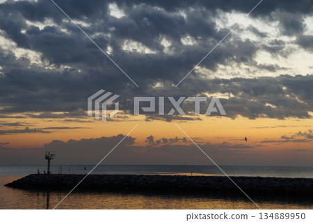 The first rays of sun paint the gray sky orange. The gray and orange sky is reflected in the sea, against the backdrop of a stone pier  134889950