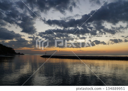 The dawn is breaking over the horizon.The first rays of sun paint the gray sky orange. The gray and orange sky is reflected in the sea, against the backdrop of a stone pier The dawn is breaking over the horizon.The first rays of sun paint the gray sky orange. The gray and orange sky is reflected in the sea, against the backdrop of a stone pier 134889951