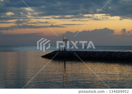 The first rays of sun paint the gray sky orange. The gray and orange sky is reflected in the sea, against the backdrop of a stone pier.The dawn is breaking over the horizon. The first rays of sun paint the gray sky orange. The gray and orange sky is reflected in the sea, against the backdrop of a stone pier.The dawn is breaking over the horizon. 134889952
