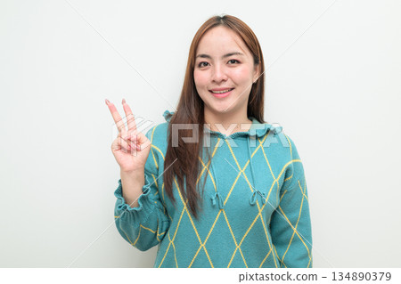 Portrait of a young Asian woman against white background making peace sign gesture Portrait of a young Asian woman against white background making peace sign gesture 134890379