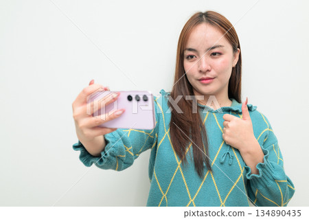 Portrait of a young Asian woman against white background using phone taking selfie 134890435