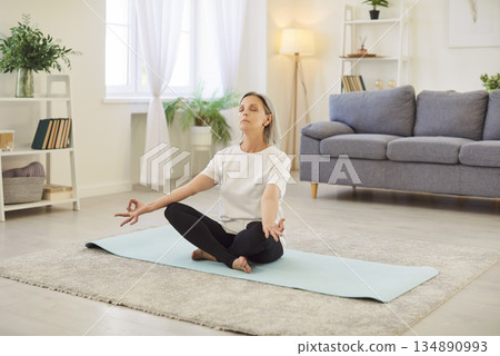 Mature woman sitting cross legged on mat, meditating with closed eyes, doing breathing exercises. Mature woman sitting cross legged on mat, meditating with closed eyes, doing breathing exercises. 134890993