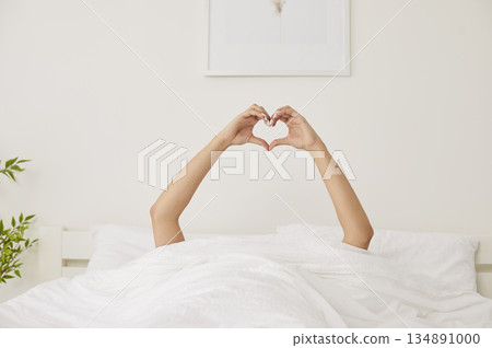 Happy young woman lying in bed, showing heart shape symbol with hands, waking up in sunny bedroom. 134891000