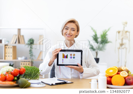 A smiling nutritionist presents a tablet with a health chart, surrounded by fresh fruits and vegetables, promoting healthy eating habits. 134892077