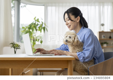 A middle-aged woman working on a computer while holding her pet dog. Image of working from home. A middle-aged woman working on a computer while holding her pet dog. Image of working from home. 134893804