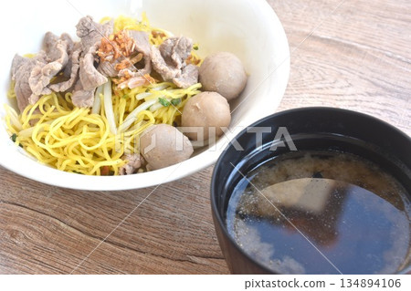 dry boiled Chinese yellow egg noodles with slice beef and meatball topping fried chop garlic on bowl with clear soup cup dry boiled Chinese yellow egg noodles with slice beef and meatball topping fried chop garlic on bowl with clear soup cup 134894106