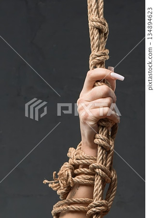 Close-up of a woman's hand with a tattoo tightly gripping a thick bondage rope against a gray background 134894623