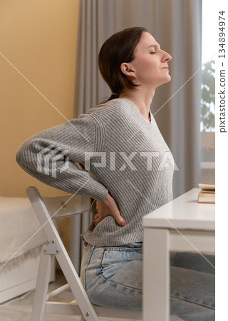 Side view unhappy young Caucasian woman suffering from backache after long hours sedentary work, sitting in home office, frustrated upset female employee worker feeling pain, touching relieving back 134894974
