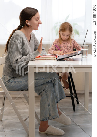 Positive Caucasian woman having an online meeting with earphones at home, sitting at a table with a laptop, while young girl draw in the background. Videocall class lecture using modern technology 134894976