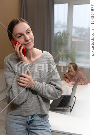 Smiling Caucasian woman talking on smartphone at home, while young girl draws at a table with a laptop in cozy living room. Young business woman freelancer speak on mobile phone behind notebook Smiling Caucasian woman talking on smartphone at home, while young girl draws at a table with a laptop in cozy living room. Young business woman freelancer speak on mobile phone behind notebook 134894977