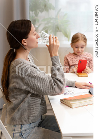 Focused Caucasian woman in gray sweater drinking water at table while young girl with smartphone sits nearby in cozy home environment. Young female sit at desk hold glass of water work from home 134894978