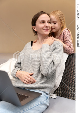 Joyful Caucasian woman sitting with a laptop, smiling while a young girl hugs her from behind, showing affection and closeness in a cozy indoor setting, playing entertaining time together at home Joyful Caucasian woman sitting with a laptop, smiling while a young girl hugs her from behind, showing affection and closeness in a cozy indoor setting, playing entertaining time together at home 134894987