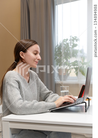 Cheerful young Caucasian woman typing on laptop, resting at home using computer for Internet communication. Freelancer girl working on comfortable desk, using service for remote job E-learning student Cheerful young Caucasian woman typing on laptop, resting at home using computer for Internet communication. Freelancer girl working on comfortable desk, using service for remote job E-learning student 134894988