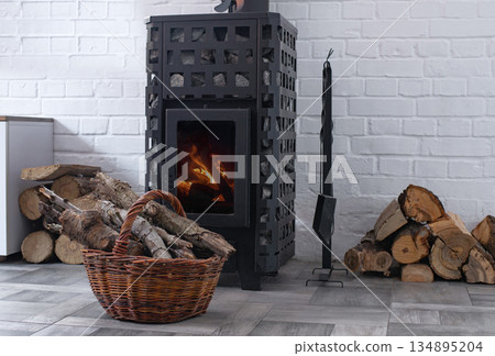 Modern Wood Stove with visible burning fire surrounded by stacked firewood and wicker log basket in a cozy Scandinavian style interior. Home heating, winter comfort and sustainable energy concept Modern Wood Stove with visible burning fire surrounded by stacked firewood and wicker log basket in a cozy Scandinavian style interior. Home heating, winter comfort and sustainable energy concept 134895204