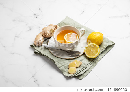 A cup of hot tea with fresh lemon and ginger on a napkin on a white marble background 134896030