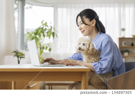 一位中年婦女一邊用電腦工作，一邊抱著她的寵物狗。這是在家工作的場景。 134897305