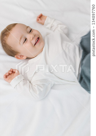 Cozy portrait of a happy baby lying on a soft white bed at home, smiling in natural light. A warm and cozy childhood moment filled with joy, innocence, comfort, and gentle family emotion. 134897760