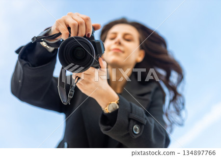 Low-angle view of a female photographer holding a professional camera against the blue sky, capturing outdoor moments and focusing on creative photography. 134897766