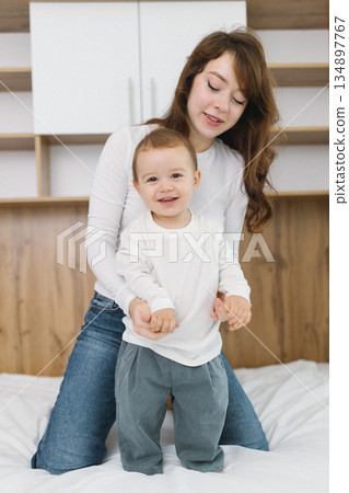 Young mother playing with her baby at home, enjoying a warm and cozy family moment full of tenderness, comfort, natural light, and playful parent-child connection. 134897767