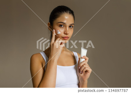 Young woman applying face cream while holding a cosmetic tube, demonstrating a hydrating skincare step on a clean studio background. Young woman applying face cream while holding a cosmetic tube, demonstrating a hydrating skincare step on a clean studio background. 134897787