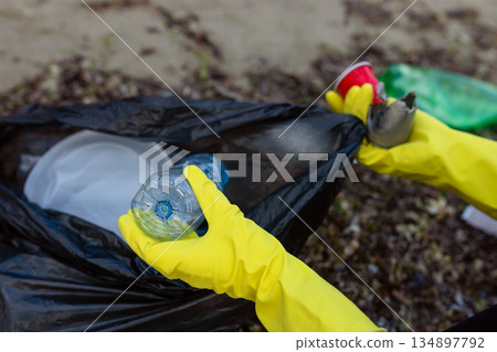 Volunteer collecting plastic waste and litter into a black trash bag during beach cleanup, highlighting pollution, environmental protection, sustainability, and eco awareness. Volunteer collecting plastic waste and litter into a black trash bag during beach cleanup, highlighting pollution, environmental protection, sustainability, and eco awareness. 134897792
