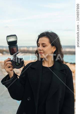 Young female photographer taking pictures outdoors by the sea, smiling while holding a professional camera with flash, capturing moments on a rocky coastline at sunset. 134897800