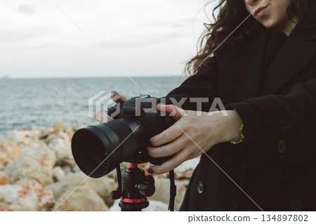 Female photographer adjusting professional camera on tripod outdoors by the sea, focusing the lens while preparing for a creative photo shoot on a rocky coastline. 134897802