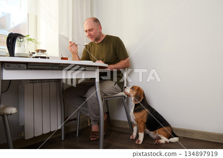 A man eats breakfast at the kitchen table, eating from a bowl while his dog sits nearby. A man eats breakfast at the kitchen table, eating from a bowl while his dog sits nearby. 134897919