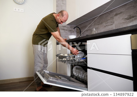 Man loading dishes into a dishwasher in a modern kitchen while standing on a wooden floor Man loading dishes into a dishwasher in a modern kitchen while standing on a wooden floor 134897922