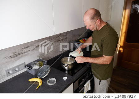 A man prepares breakfast, pours milk into a saucepan, and cooks porridge on the stove. A man prepares breakfast, pours milk into a saucepan, and cooks porridge on the stove. 134897927