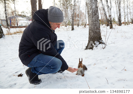 A man sits by a tree in a snowy forest. He reaches out to feed a squirrel some nuts. The squirrel perches on his hand. 134898177