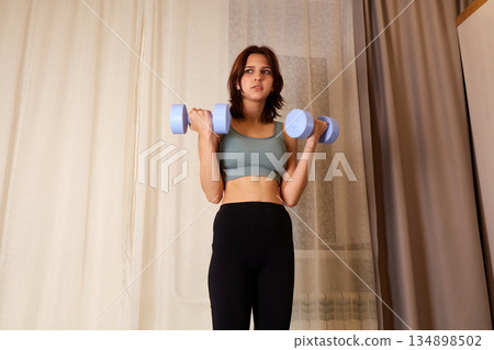 Young woman in sportswear is lifting dumbbells while standing in a home gym. . Young woman in sportswear is lifting dumbbells while standing in a home gym. . 134898502