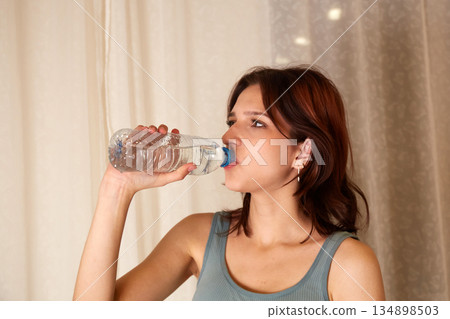A woman holds a water bottle and drinks from it while standing indoors. A woman holds a water bottle and drinks from it while standing indoors. 134898503