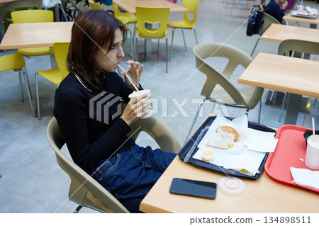 Woman is seated in a food court, sipping a drink from a cup. 134898511